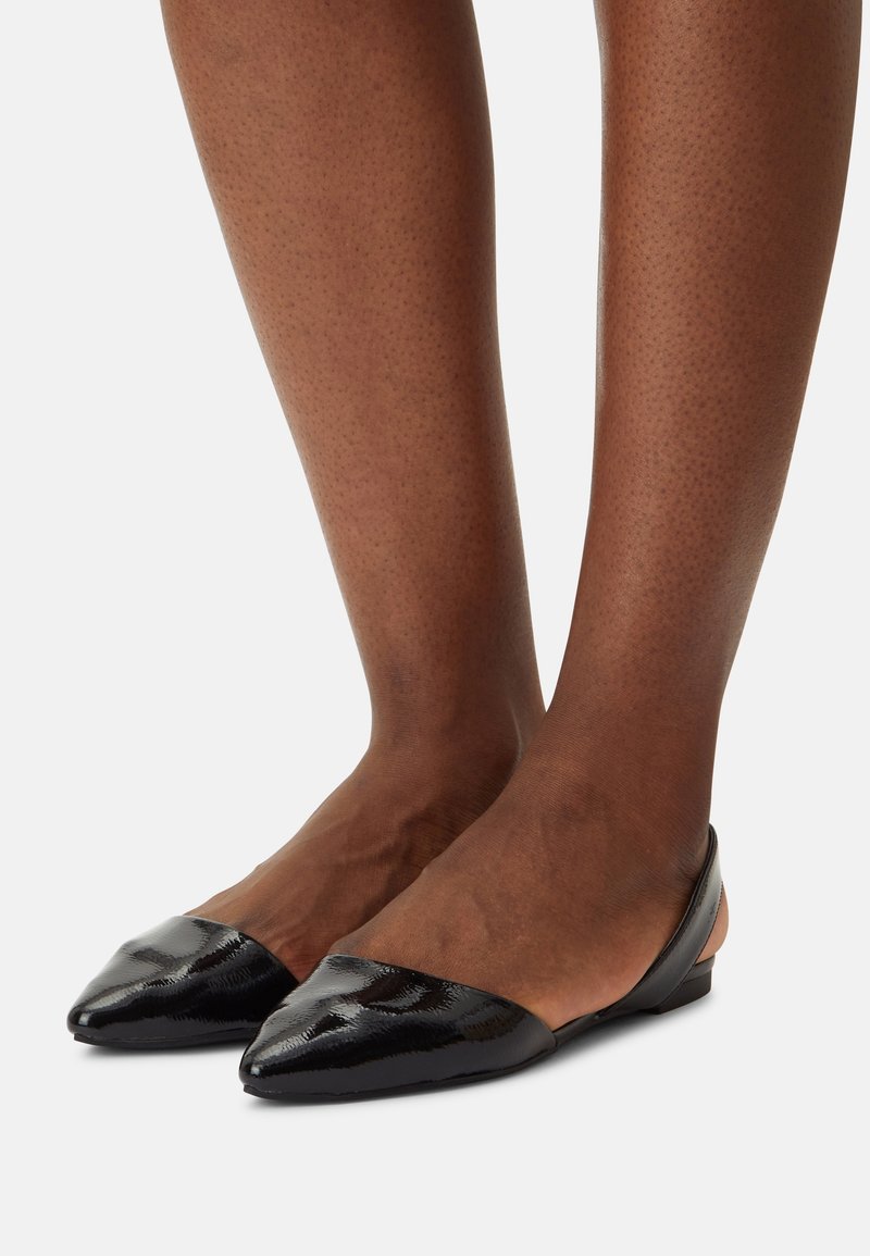Jambes à peau foncée portant des ballerines slingback noires en cuir verni à bout pointu avec un petit talon sur un fond blanc.