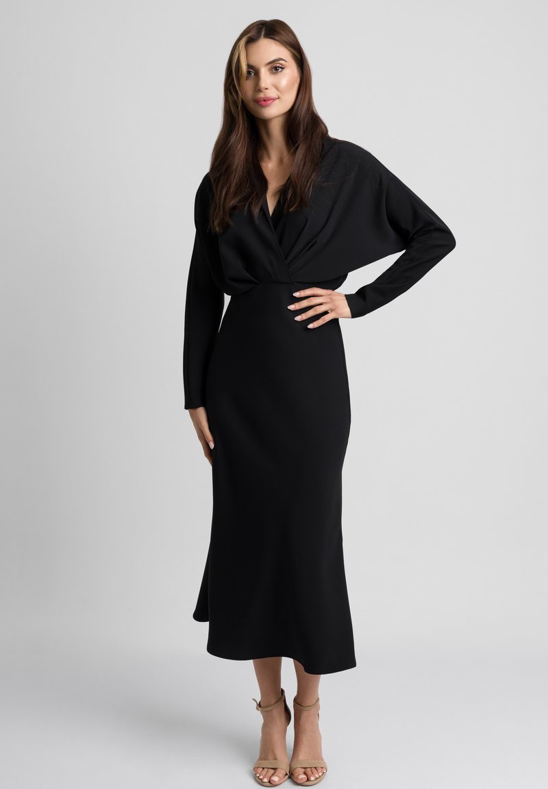 Długa czarna sukienka z wiązanym gorsetem, długimi rękawami i gładką teksturą. W połączeniu z jasnobeżowymi sandałami na wysokim obcasie.