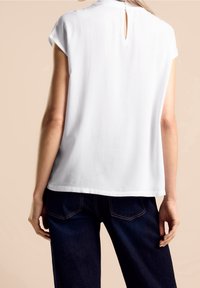 Blusa bianca a maniche corte con scollatura rotonda, caratterizzata da un'apertura a goccia sulla schiena, realizzata in un tessuto liscio e leggero.
