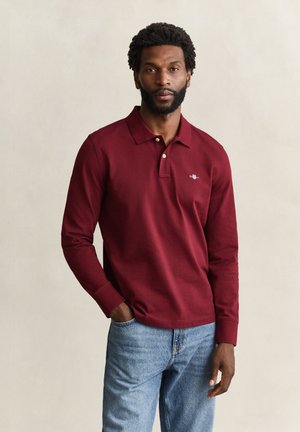 Homme avec barbe et cheveux bouclés porte un polo rouge à manches longues et un jean bleu clair, debout avec une main dans la poche.