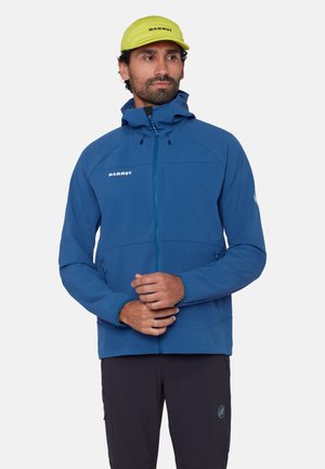 Mann trägt eine blaue Kapuzenjacke und eine neongelbe Mütze, steht mit verschränkten Händen und schaut seitlich vor einem einfarbigen Hintergrund.