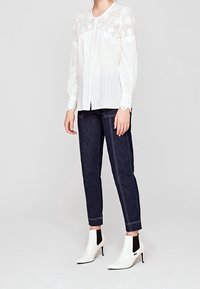 Witte blouse met lange mouwen en kantdetails, met een voorpasje, gecombineerd met donkere jeans en witte enkellaarsjes met zwarte accenten.