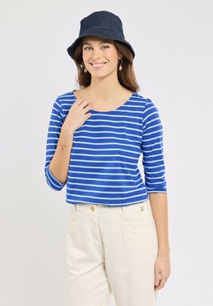 Femme portant un haut rayé bleu et blanc à manches trois-quarts, un pantalon crème taille haute, un chapeau seau sombre, et des boucles d'oreilles créoles dorées, souriante.