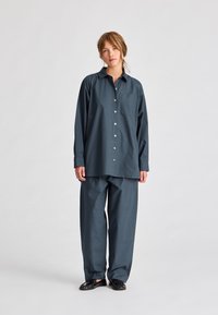 Tmavě modrá, volná košile s dlouhými rukávy a přední kapsou, spárovaná s odpovídajícími širokými kalhotami. Tkanina vypadá měkce a hladce.