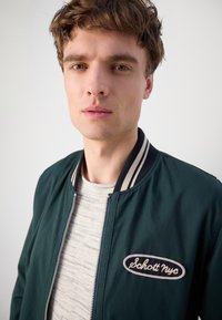 Chaqueta bomber verde con un collar acanalado a rayas, cierre de cremallera y parche bordado de "Schott NYC" en el pecho, llevada sobre un suéter claro con estampado.