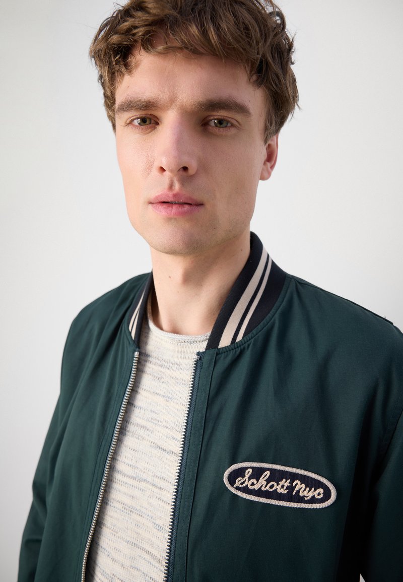 Veste bombardier verte avec un col côtelé rayé, fermeture éclair et écusson brodé "Schott NYC" sur la poitrine, portée sur un pull léger à motifs.