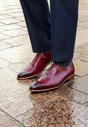 Bordeaux leren veterschoenen met een slank, puntig ontwerp en een gladde textuur, gecombineerd met donkere broeken op een natte, stenen ondergrond.