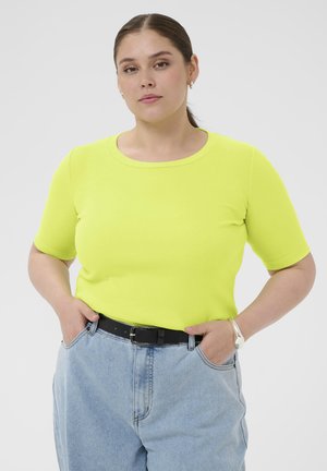 Ung kvinde med tilbagelagt hår iført en knaldgul kortærmet top og lyseblå højhælede jeans med hænderne i lommerne.