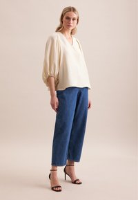 Blusa di lino crema con scollatura a V e maniche a palloncino, abbinata a pantaloni di jeans blu a vita alta. Modella che indossa tacchi neri con cinturini.