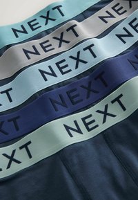 Ceintures empilées de shorts bleu marine avec des bandes élastiques dans diverses nuances de bleu et de gris, chacune étiquetée "NEXT" en lettres noires grasses.