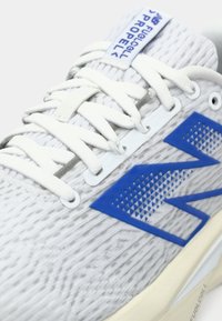 Chaussure de sport grise avec un tissu texturé, des lacets blancs et des accents bleus, présentant un motif géométrique sur le côté et une semelle rembourrée.