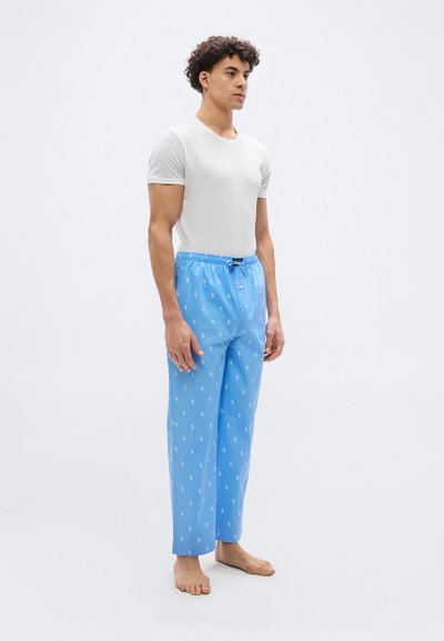 Νέος άνδρας στέκεται ξυπόλητος, φορώντας λευκό T-shirt και γαλάζιες πιτζάμες με μικ ρά λευκά λογότυπα σε απλό φόντο.