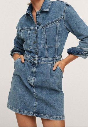 Femme portant une robe en jean bleue boutonnée, avec de longues manches retroussées et les mains dans les poches avant.