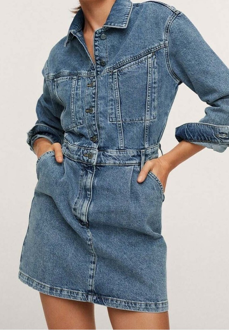 Femme portant une robe en jean bleue boutonnée, avec de longues manches retroussées et les mains dans les poches avant.