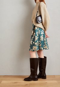 Beige, gerippter Pullover, tailliert, mit abgesenkten Schultern; grüner, floral bedruckter Rock, oberhalb des Knies; hohe, dunkelbraune Lederstiefel mit niedrigen Absätzen.