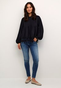 Melnas, teksturētas garām piedurknēm topa ar izplūdušu dizainu un mežģīņu apdari ap apakšmalu, apvienots ar ziliem šauriem džinsiem un beiges čības.