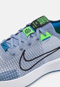 Zapatilla deportiva azul claro con parte superior de malla, logo de Nike en negro, detalles de acento en verde y una suela con textura patrón. Incluye una etiqueta con código QR.