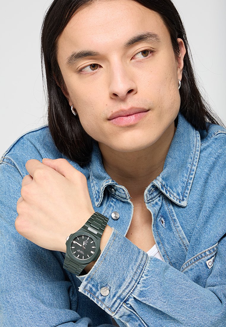 Reloj de pulsera de plástico verde con una correa texturizada, esfera negra, marcadores de horas blancos y acentos plateados, llevado en una muñeca vestida con chaqueta de mezclilla.