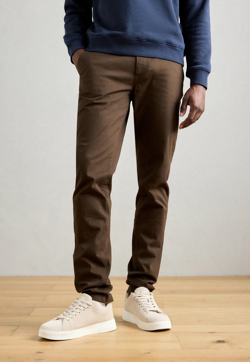Brown cotton pants with a slim fit, featuring a smooth texture. Paired with cream-colored suede sneakers, set against a wooden floor.