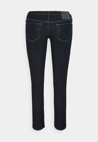 Dunkelblaue Jeans mit einer taillierten Passform, hohem Bund, gelben Nähten und zwei Gesäßtaschen. Mit einem Markenpatch am Bund.