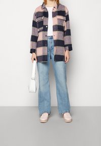 Osoba w dużej, wzorzystej kurtce w różowe, czarne i fioletowe kratki, jasnoniebieskich prostych dżinsach, różowych sneakersach, trzymająca małą białą torebkę.