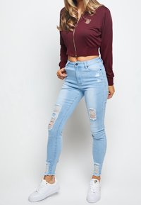 Bordowa krótka kurtka z zamkiem, wykonana z teksturowanej tkaniny i z złotym logo; jasnoniebieskie wysokie dżinsy z przetarciami; białe sneakersy z złotymi akcentami.