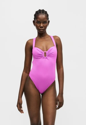 Kvinde med flettet hår iført en ærmeløs, pink heldragt med en nøglehul-udskæring ved brystet, stående mod en ensfarvet baggrund.