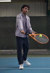Sportaš u svijetloplavoj jakni i odgovarajućoj majici, drži narančasti teniski reket s zelenom lopticom, nosi tamne hlače i bijele tenisice.
