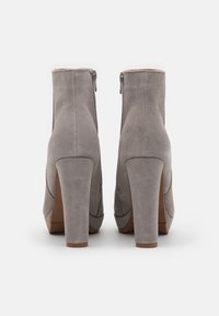 Par de botas de ante gris hasta el tobillo con tacones altos y gruesos y cremalleras en la parte trasera, mostradas desde atrás sobre un fondo blanco.