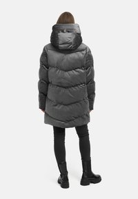 Persona, kas uzvilkusi tumši pelēku pārspīlētu pūkaini mēteli ar lielu kapuci, melnas pieguļošas bikses un melnas masīvas potīšu zābakus, stāv ar muguru uz skatītāju.