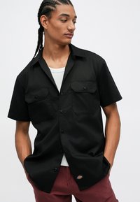 Homem com cabelo trançado, vestindo uma camisa preta de manga curta de botões sobre uma t-shirt branca e calças em tom borgonha, olhando para o lado com as mãos nos bolsos.