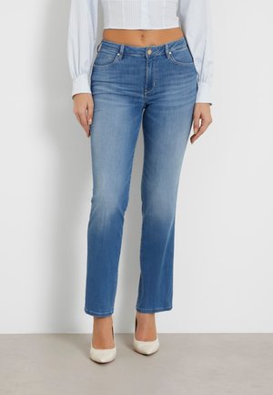 Kvinde iført lyseblå jeans med lige ben, cropped hvid langærmet skjorte og hvide spidse stilethæle, stående mod en hvid væg.