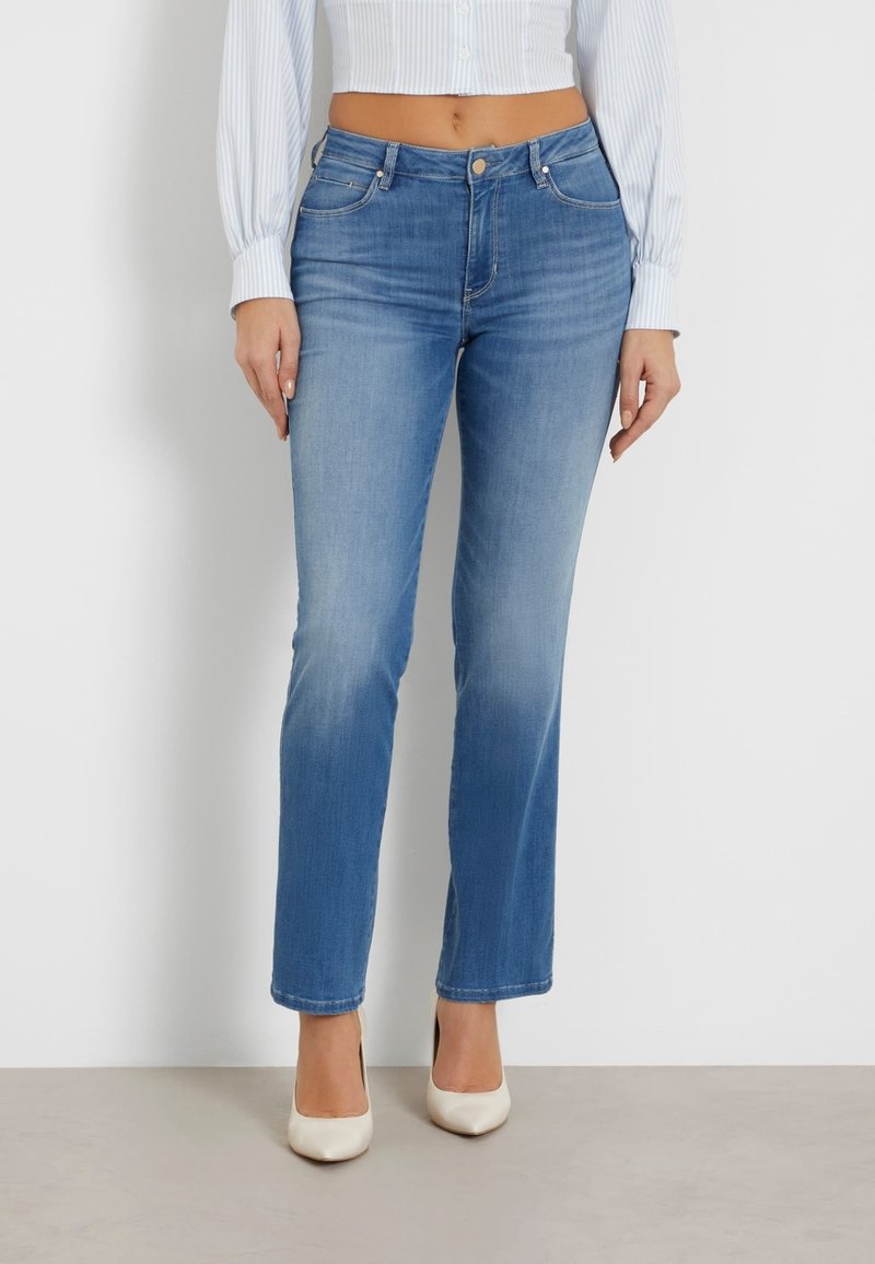 Femme portant un jean bleu clair droit, un haut blanc court à manches longues et des talons blancs pointus, debout contre un mur blanc.