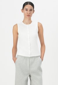 White sleeveless top with a round neckline and button closure down the front. Smooth texture with a slim fit. Paired with gray sweatpants.