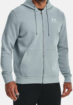 Homme portant un sweat à capuche gris clair avec fermeture éclair, poches avant et logo Under Armour, associé à un pantalon noir.
