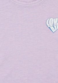 Camiseta lila de tela suave con un pequeño diseño de corazón azul y el texto "AMOR EN EL AIRE" en una fuente estilizada.