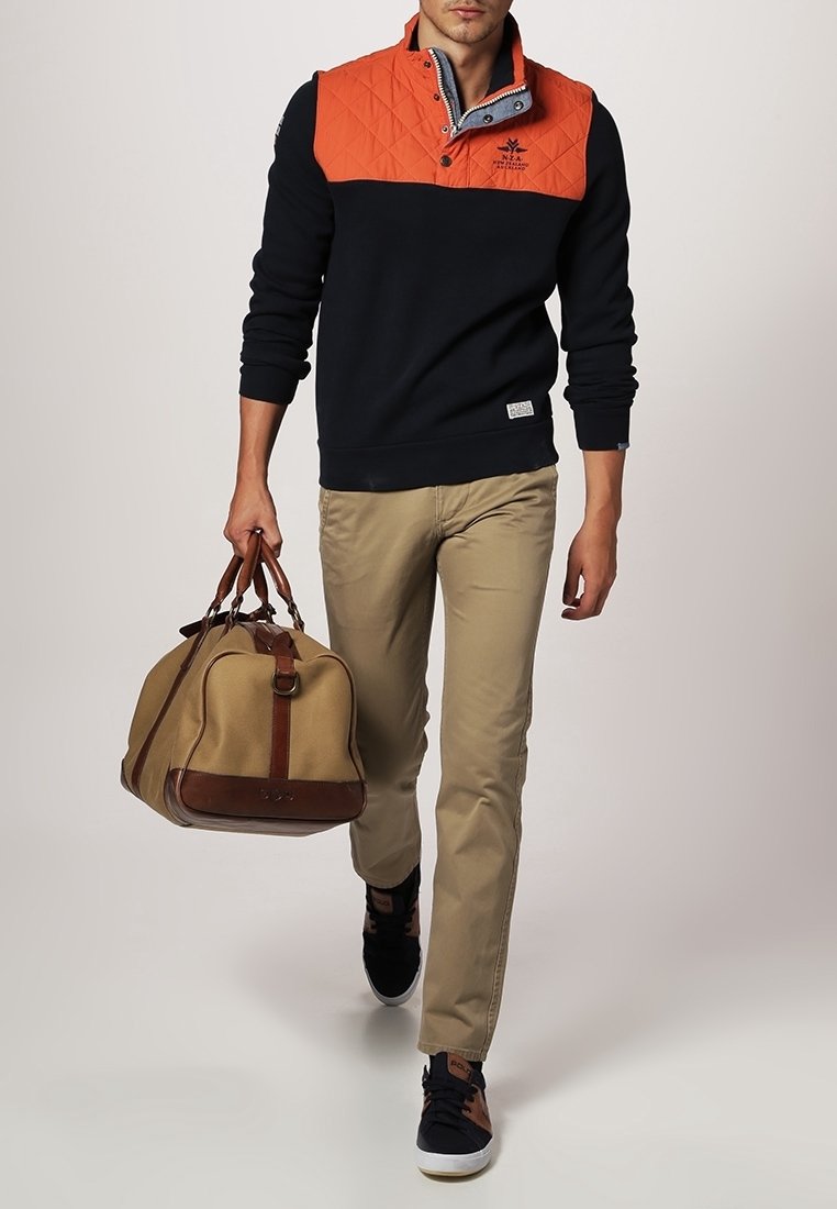 Homme marchant en pantalon beige, pull noir et orange, baskets noires, portant un sac de voyage en cuir beige et marron dans la main droite.