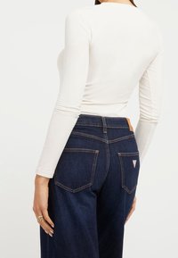 Top bianco a maniche lunghe abbinato a jeans a vita alta blu scuro con due tasche posteriori e una toppa in pelle in vita.