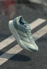 Ανοιχτό μπλε παπούτσι Adidas για τρέξιμο με λευκές ρίγες, κρεμασμένο πάνω από την άσφαλτο με λευκές λωρίδες οδού.