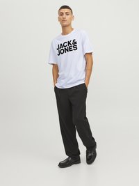 Balts īsām piedurknēm T-krekls ar izteiksmīgu melnu "JACK & JONES" tekstu, kombinēts ar brīvām melnām biksēm un melnām kurpēm, stāvot uz neitrāla fona.