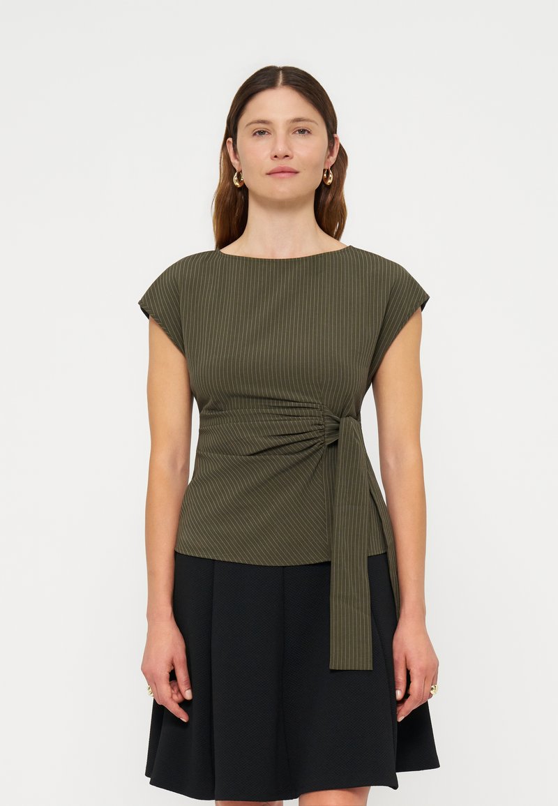 Femme portant un haut vert olive à fines rayures avec détail noué sur le côté et une jupe noire, debout sur un fond blanc uni.