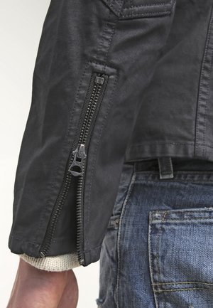 Schwarzer Jackenärmel mit teilweise geöffnetem vertikalem Reißverschluss über der Tasche einer blauen Denim-Jeans und cremefarbenem Strickbündchen am Handgelenk sichtbar.