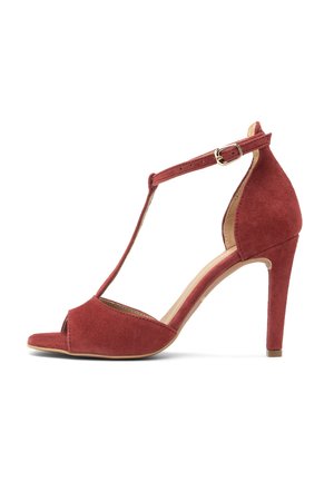 Czerwona zamszowa sandałka na wysokim obcasie z paskiem w kształcie litery T, odkrytymi palcami, regulowaną klamrą przy kostce i smukłym obcasem na jednolitym białym tle.