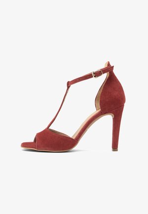 Czerwona zamszowa sandałka na wysokim obcasie z paskiem w kształcie litery T, odkrytymi palcami, regulowaną klamrą przy kostce i smukłym obcasem na jednolitym białym tle.