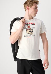 Camiseta de color crema con un diseño gráfico de una calavera, una mujer y rosas. Lleva puesta una chaqueta de cuero negra y pantalones negros.