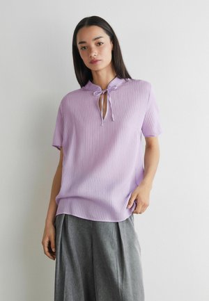 Junge Frau mit langen dunklen Haaren, die ein fliederfarbenes, geripptes Kurzarmshirt mit Neckholder und lockere graue Hose trägt, steht vor einer weißen Wand.