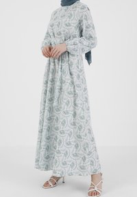 Pitkä, ilmava mekko kevyestä kankaasta, jossa on vihreä lehväkuvio valkoisella taustalla. Mekossa on puffipuffit ja vyötärö on kiristetty.