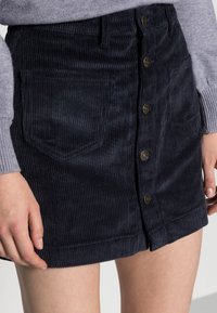 Marinblå kordflöjel mini kjol med framknäppning, två sidofickor och vertikal ribbad textur. Nederkant slutar vid mitten av låret.