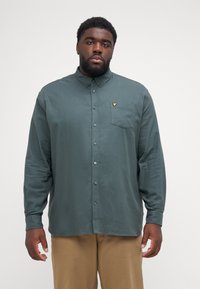 Camisa de manga larga verde oscuro con cuello abotonado, tejido texturizado, un bolsillo en el pecho y un pequeño logotipo amarillo en el bolsillo.