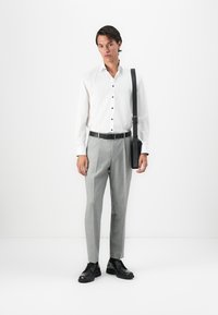 Man wearing white buttoned shirt, gray tailored pants, black belt, black leather shoes, and carrying a black shoulder bag on white background.